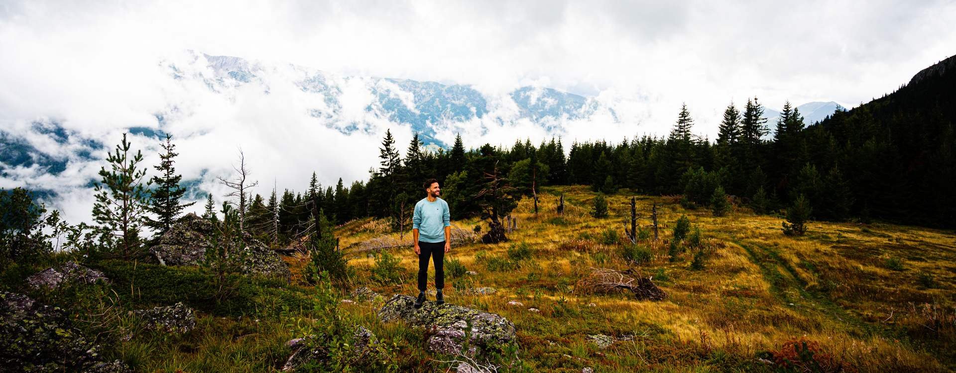 Man staat op een rots in Albanië met uitzicht op een bergachtig, bosrijk landschap en wolken boven de verre toppen