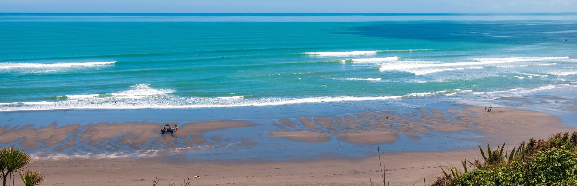 Turquoise oceaangolven rollen het zandstrand op waar een kleine tractor en mensen aanwezig zijn aan de kust