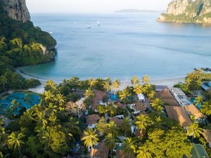 Luchtfoto van een strand resort aan een rustige baai, omringd door palmbomen en groen. Op de achtergrond liggen enkele zeilboten op het kalme, blauwe water.