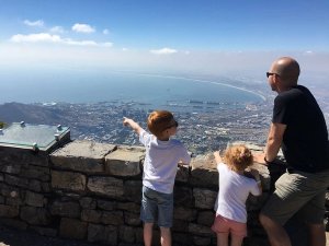 Man met twee jonge kinderen staat op een berg en kijkt uit over een stad, haven en baai, terwijl één kind wijst in Zuid-Afrika