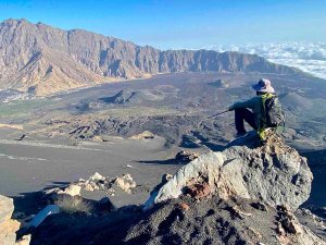 Persoon zit op een rotsachtige bergtop en kijkt uit over een uitgestrekte vulkanische caldera met donkere asvlaktes en kleinere vulkaankegels in een vulkanisch natuurgebied