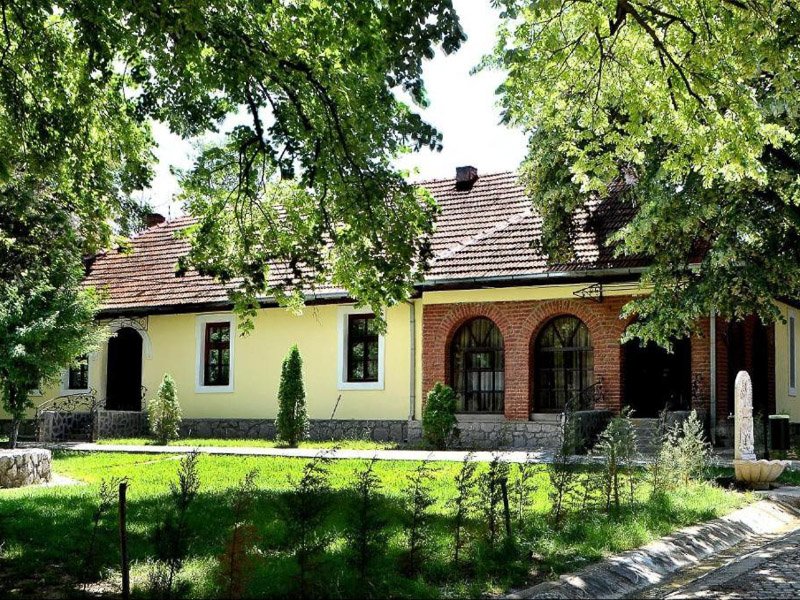 Laag gebouw in Kosovo met gele en bakstenen gevel met boogramen, omringd door gras en bomen
