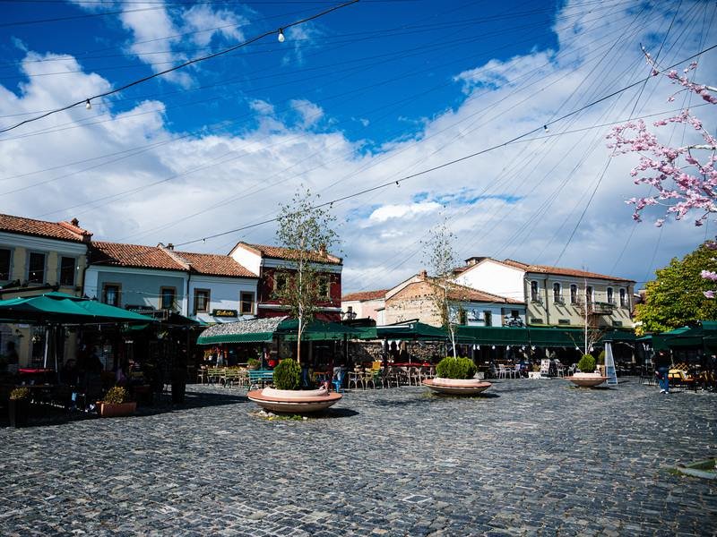 Levendig plein in Albanië met geplaveide straat, historische gebouwen, groene marktkramen, bomen en lichtslingers onder een blauwe lucht