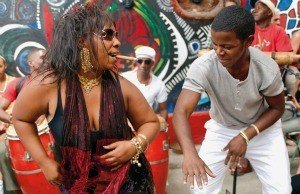 Vrouw met gouden sieraden en een man dansen energiek buiten voor een kleurrijke muurschildering en trommels in Cuba, Havana