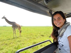 Vrouw glimlacht terwijl ze vanuit een safarivoertuig uitkijkt over de savanne, met een giraffe op de achtergrond