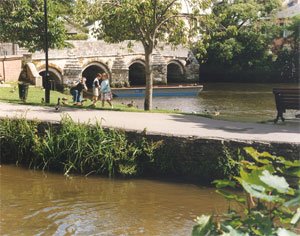 Stenen boogbrug overspant een rivier met mensen en eenden op de grasrijke oever en een blauwe boot op het water in een historische stad