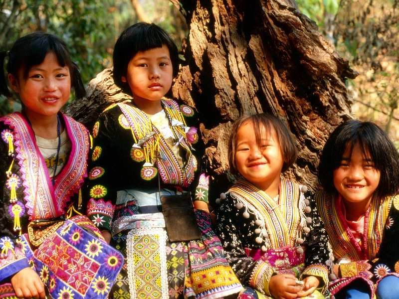 Vier Hmong-meisjes in traditionele kleurrijke kleding zittend bij een boom.