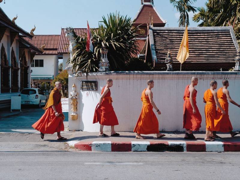 Zeven mannen in oranje gewaden lopen op blote voeten over een trottoir langs een witgekalkte muur. Op de achtergrond staan traditionele gebouwen met puntdaken en decoratieve elementen in Chiang Mai