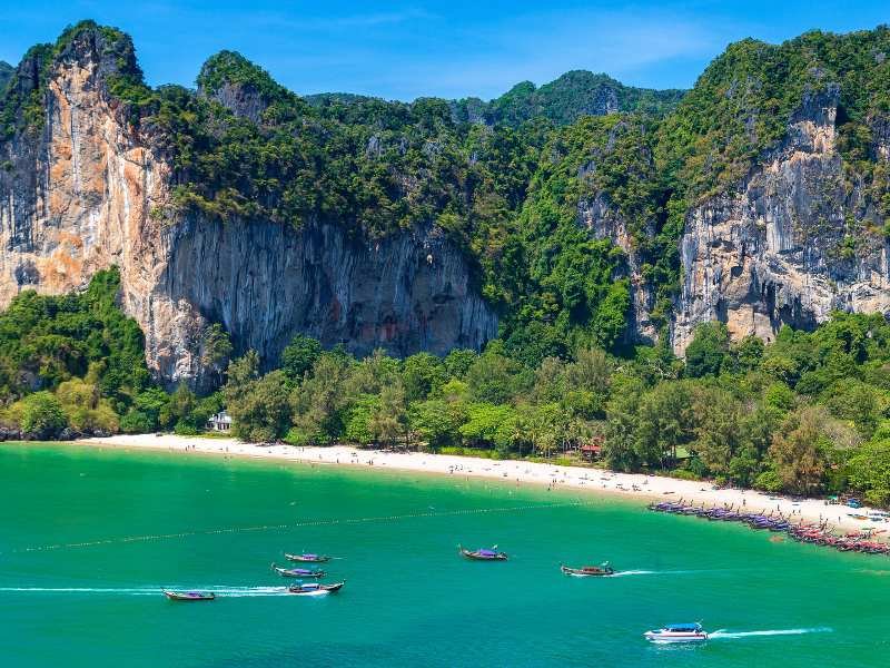 Uitzicht op een tropisch strand aan helder turquoise water, omringd door dichtbegroeide jungle. Op de achtergrond torenen steile kalkstenen kliffen omhoog, deels begroeid met groen. Op zee varen traditionele longtailboten en een enkele speedboot, terwijl mensen op het strand ontspannen of wandelen.