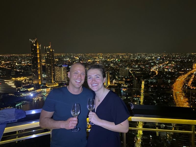 Twee mensen met een glas wijn in de hand poseren op een rooftopbar met uitzicht op een grote verlichte stad in de avond, met hoge gebouwen en een kronkelende snelweg op de achtergrond. Het is donker buiten.
