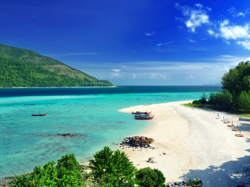 Tropisch wit zandstrand met turquoise zee, longtailboten en groene heuvels onder een heldere blauwe lucht.