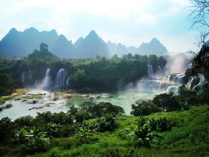 Uitzicht op de Ban Gioc-waterval met meerdere watervallen die uitmonden in een turquoise rivier, omringd door groene vegetatie en kalkstenen bergen op de achtergrond.