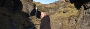 Lachende vrouw in een zwarte jas staat voor een hoge waterval die langs met mos bedekte kliffen naar beneden stroomt in IJsland