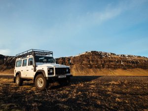 Een witte jeep staat geparkeerd in een ruig landschap met kale vlaktes en een rotsachtige bergwand op de achtergrond.