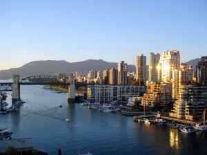Uitzicht op de skyline van Vancouver met hoge gebouwen langs het water, een brug en jachthaven, tegen een achtergrond van bergen bij zonsondergang.