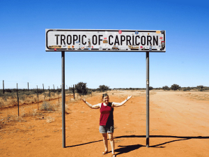 Vrouw staat met gespreide armen onder een verkeersbord met de tekst “Tropic of Capricorn” langs een droge, rode zandweg in een zonnig woestijnlandschap.