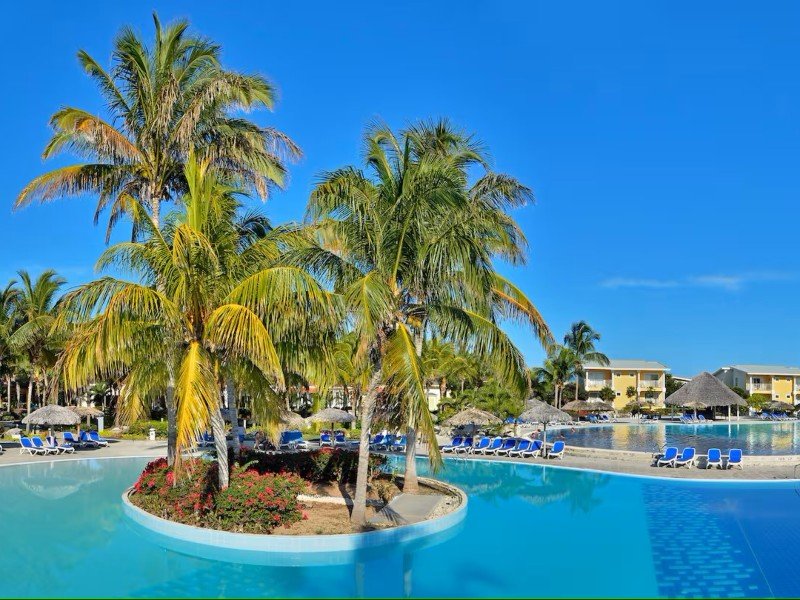 Een zonnig tropisch resort met een groot zwembad, palmbomen, ligstoelen en gele gebouwen.