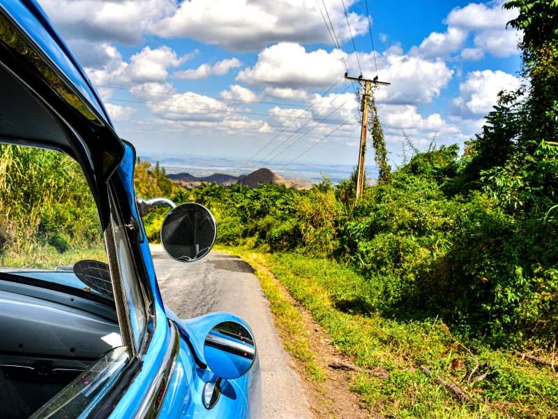 Blauwe vintage auto op een landelijke weg door een groen landschap, met een elektriciteitspaal en bergen in de verte onder een bewolkte lucht in Cuba