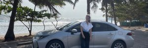 Een vrouw leunt lachend tegen een zilverkleurige huurauto, geparkeerd onder palmbomen met uitzicht op de zee. Op de achtergrond slaan de golven tegen het strand.