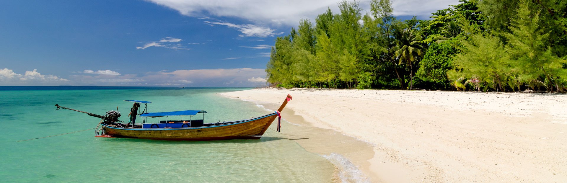 Een longtailboot in helder water naast een wit zandstrand met tropische bomen in Thailand.