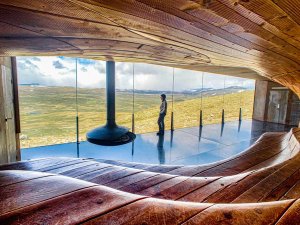 Inside a modern architectural building with a wave-like wooden interior and a suspended fireplace, a person looks out a large window at a vast, green, hilly landscape.