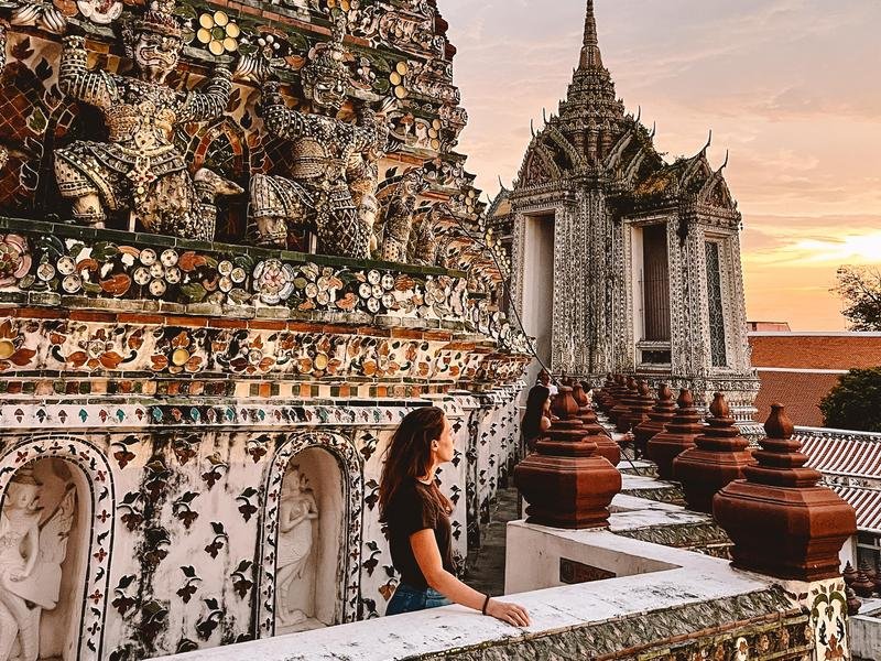 Een vrouw kijkt uit over een rijkversierde tempel in Bangkok met kleurrijke mozaïeken en beelden van wachters. Op de achtergrond is een tweede tempelgebouw te zien met een spitse toren, terwijl de lucht warm kleurt door de ondergaande zon.