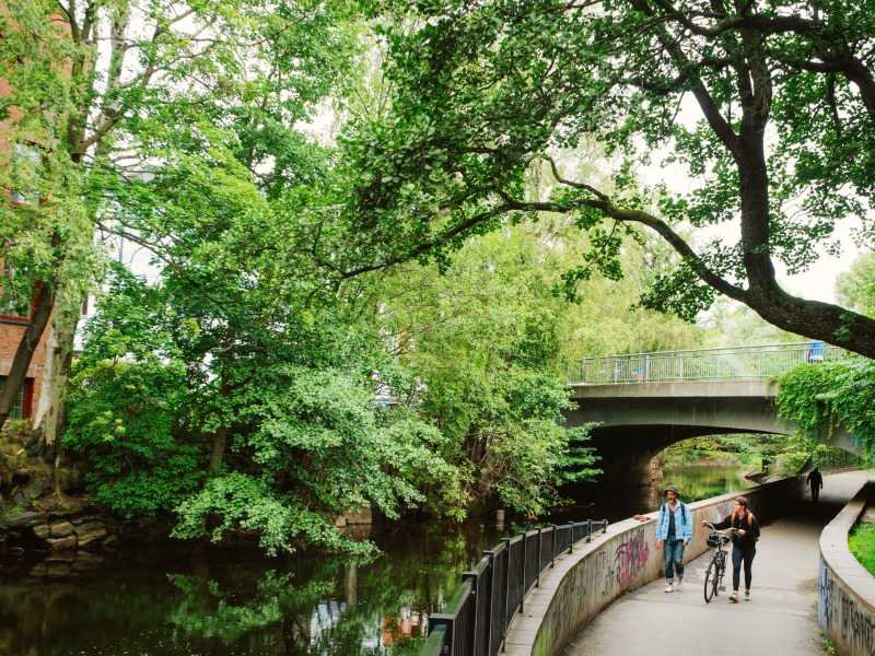 Two people walk along a path next to a tree-lined canal, with a bridge in the background.