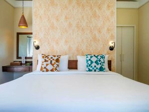 Comfortabel tweepersoonsbed met decoratieve kussens en warme verlichting, tegen een zacht patroonbehang.