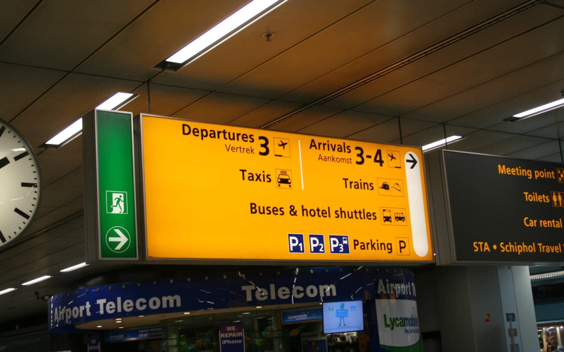 Geel informatiebord op een luchthaven met aanwijzingen voor vertrek, aankomst, taxi’s, treinen, bussen, hotelshuttles en parkeerplaatsen.