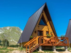 Houten A-frame cabin met donker dak en terras, met bergen onder een blauwe lucht in Kroatië