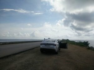 Witte auto met open deur geparkeerd op een zandberm langs een weg met uitzicht op de zee onder een bewolkte lucht in Cuba