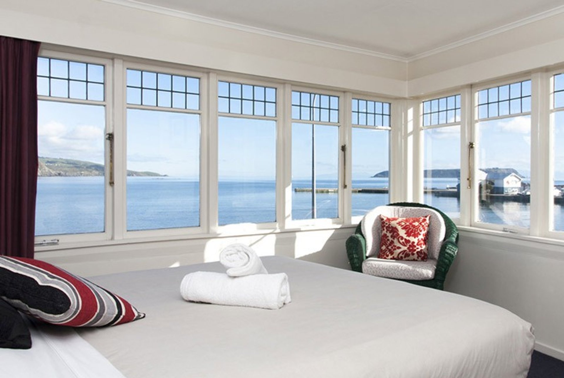 Slaapkamer met erker en uitzicht op de oceaan en een haven met een bed en rieten stoel aan de kust