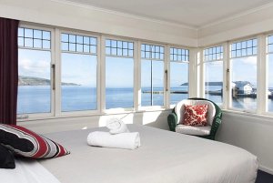 Slaapkamer met erker en uitzicht op de oceaan en een haven met een bed en rieten stoel aan de kust