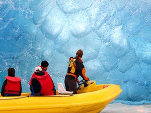 Gele boot met drie mensen in reddingsvesten vaart richting een grote blauwe ijswand in een gletsjergebied