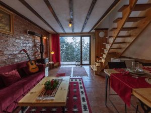 Rustieke woon- en eetkamer in Kosovo met rode bank, bakstenen muur, open haard, houten trap en groot raam met uitzicht op het bos
