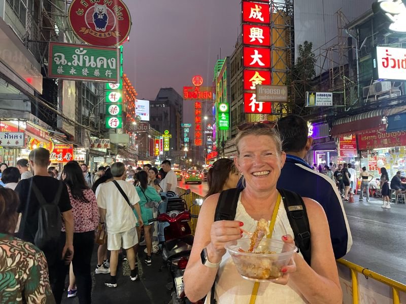 Vrouwelijke reiziger eet s'avonds streetfood in een drukke straat vol neonreclames in Bangkok’s Chinatown.