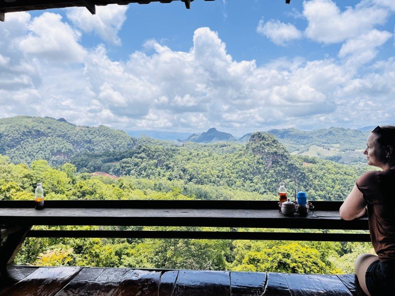 Uitzicht vanaf een terras op beboste bergen met wolken in de lucht. Een vrouwelijke reiziger zit aan een bar die richting het uitzicht geplaatst is.
