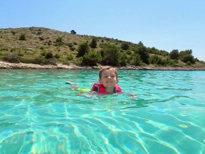 Lachende jongen in rood reddingsvest die zwemt in helder turquoise water bij een rotsachtige kust in Kroatië