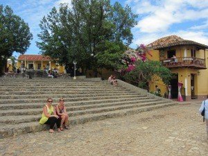 Twee vrouwen zitten op brede stenen trappen op een zonnig plein met gele gebouwen en bomen bij Trinidad in Cuba