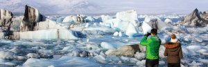 Twee mensen kijken uit over een gletsjerlagune vol ijsbergen waarbij één persoon een foto maakt in IJsland
