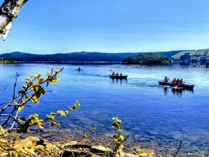 People kayaking on a calm blue lake with forested hills and an island.