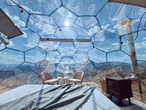 Interieur van een transparante geodetische dome met bed, twee stoelen en een kachel, met panoramisch uitzicht op een zonnig berglandschap bij Mavrovo in Noord-Macedonië