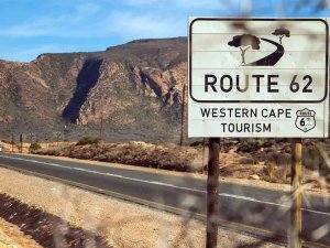 A white sign for "Route 62 Western Cape Tourism" stands next to a road in a dry, mountainous landscape.