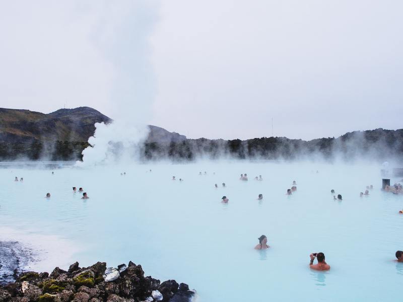 Mensen baden in een melkblauwe geothermische lagune met opstijgende stoom en heuvels in de verte bij de Blue Lagoon in IJsland