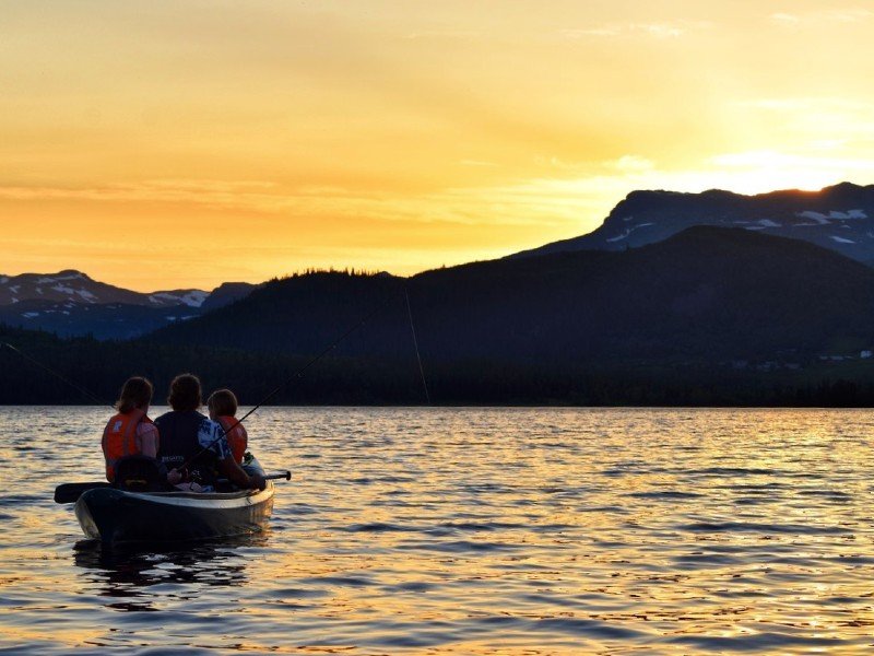 Three people fish from a canoe on a lake at sunset with mountains in the background.