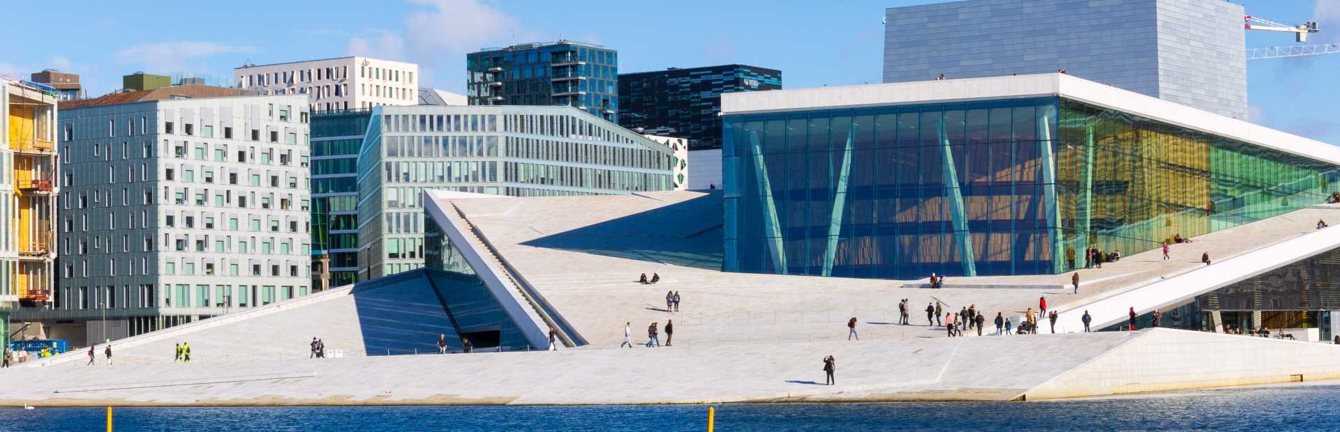 Modern Oslo Opera House with a white sloping roof, glass facade, and people walking on its surface by the water.
