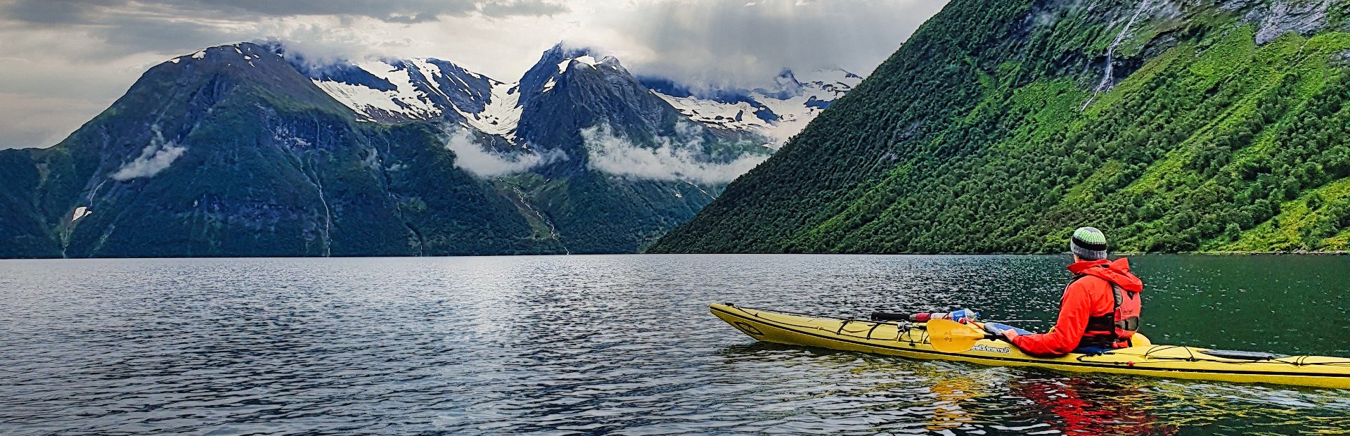 A person in a yellow kayak paddles on a serene fjord, flanked by steep green mountains and distant snow-capped peaks under a cloudy sky.