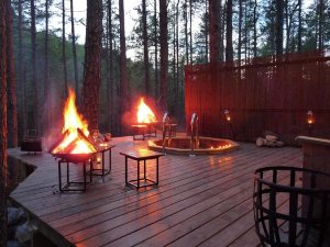 A cozy forest deck at dusk with two bonfires and a candlelit hot tub.