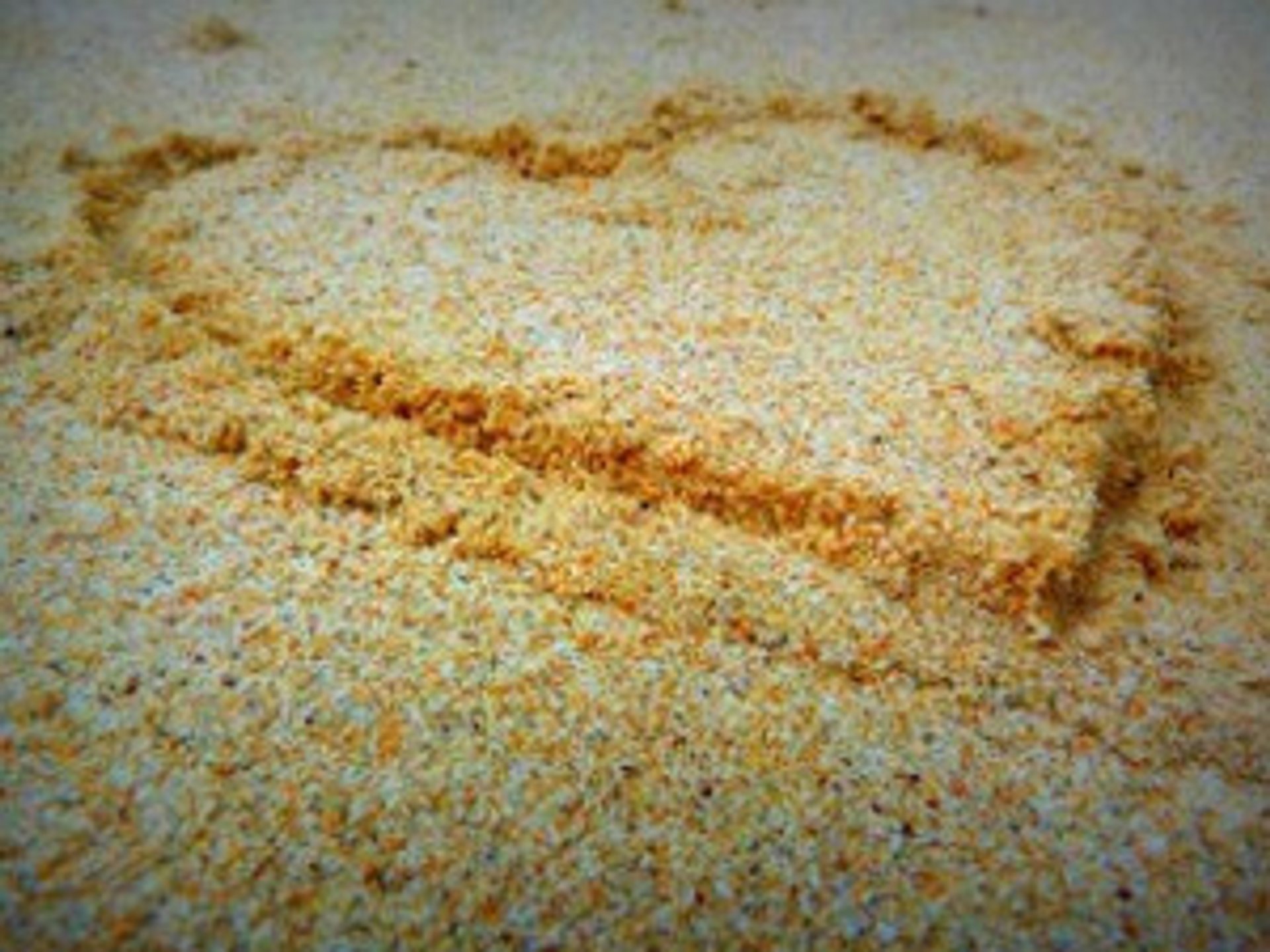 Een hartvorm is getekend in het zand op een strand. De lijnen van het hart zijn lichtjes ingedrukt in het grove, geelbruine zand.