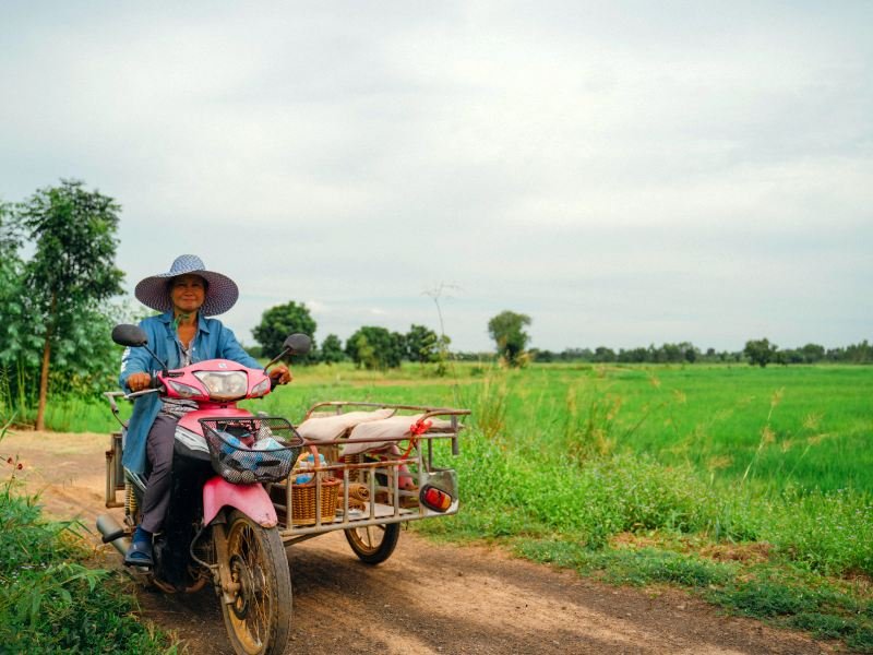 Lachende Thaise vrouw rijdt op een roze scooter met zijspan over een zandpad, omringd door groene rijstvelden en onder een bewolkte hemel.